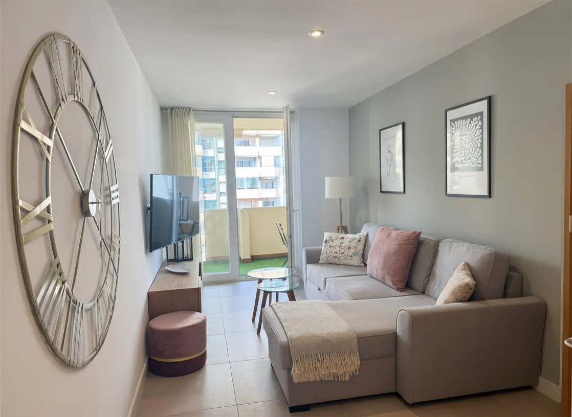 Living room with gray walls, a large clock, a TV, and a gray sectional sofa with pink pillows. A balcony is visible through sliding glass doors.