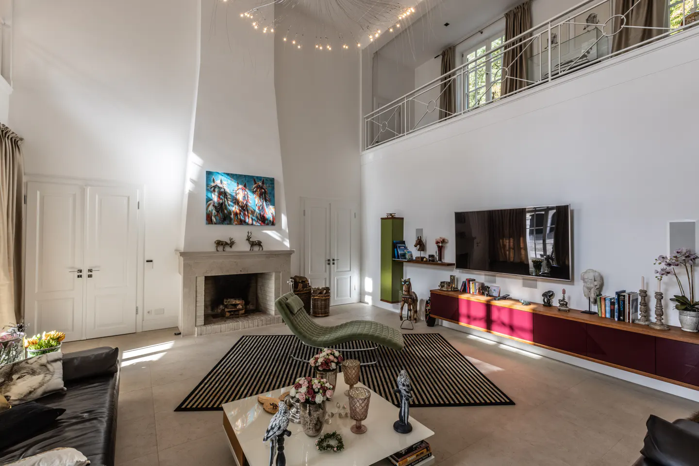 Bright, modern living room with high ceilings, a fireplace, and a second-floor balcony. A black and white striped rug sits under a green chaise lounge.