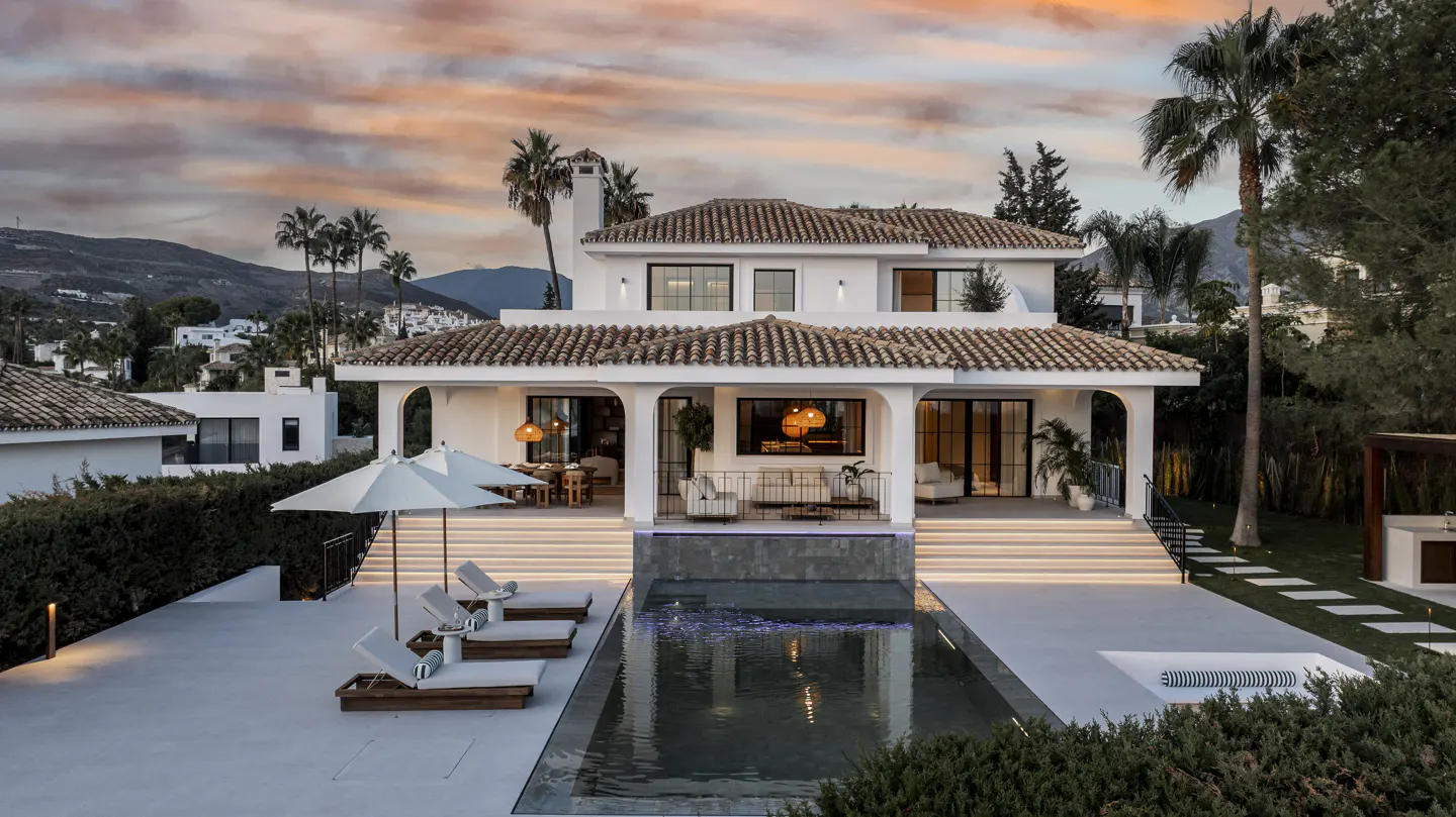 Luxury white villa with a pool at sunset. Lounge chairs and palm trees surround the property.