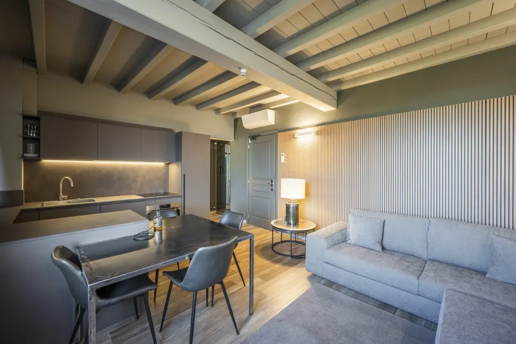 Interior view of a modern apartment with a kitchen, dining table, and gray sofa. The ceiling has exposed wooden beams.