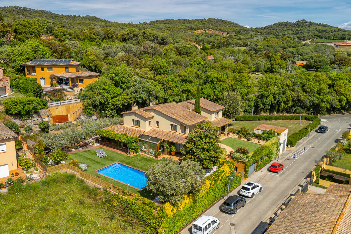 Aerial view of a tan two-story house with a blue pool, green lawn, and cars parked on the street. Trees and hills in the background.