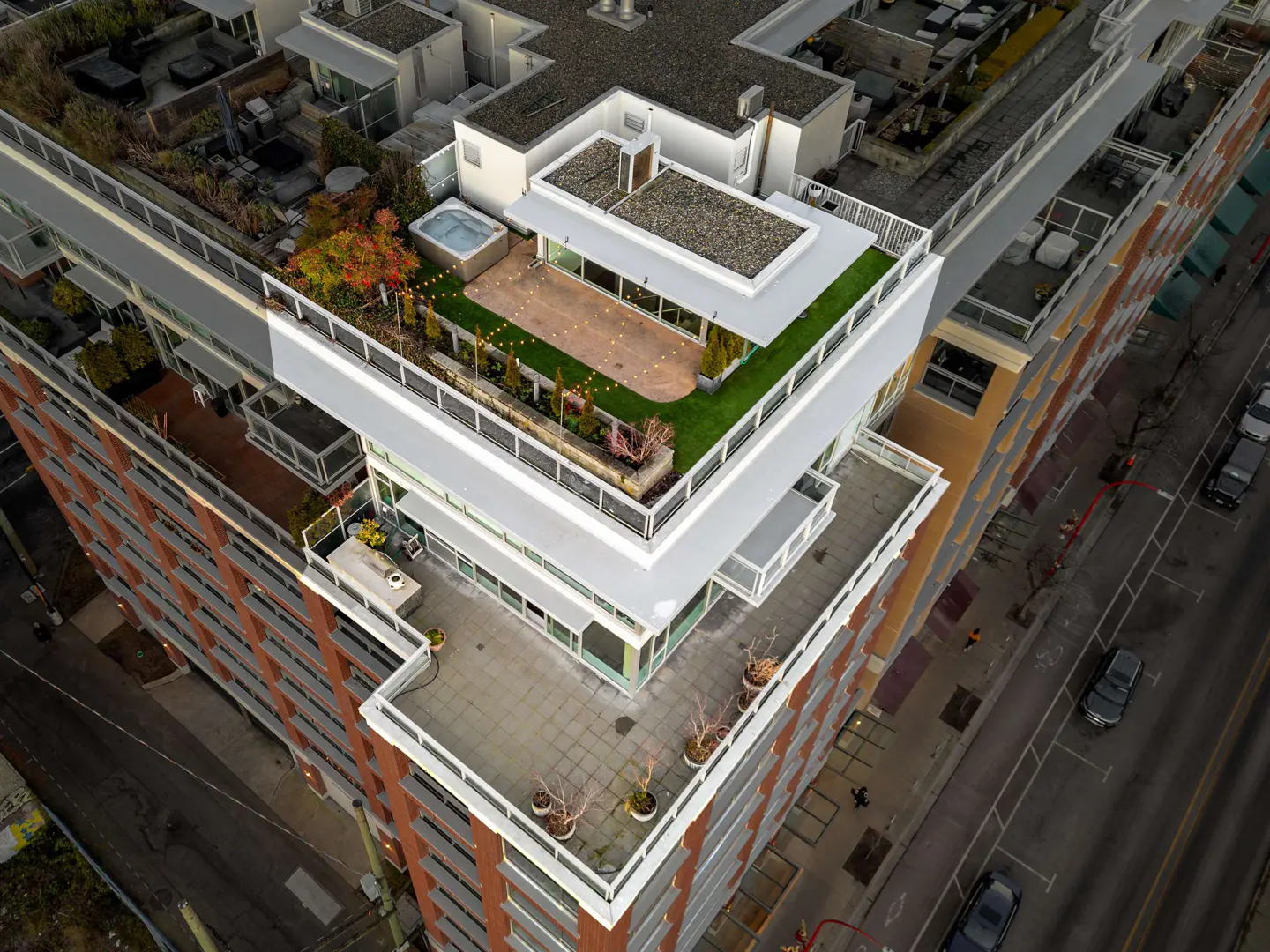 Aerial view of a modern apartment building with rooftop terrace, hot tub, string lights, and green space.