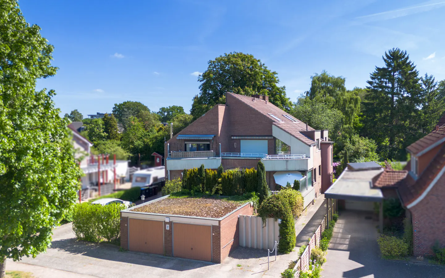 A modern brown brick house with a flat-roofed double garage, balconies, and lush green trees under a clear blue sky.