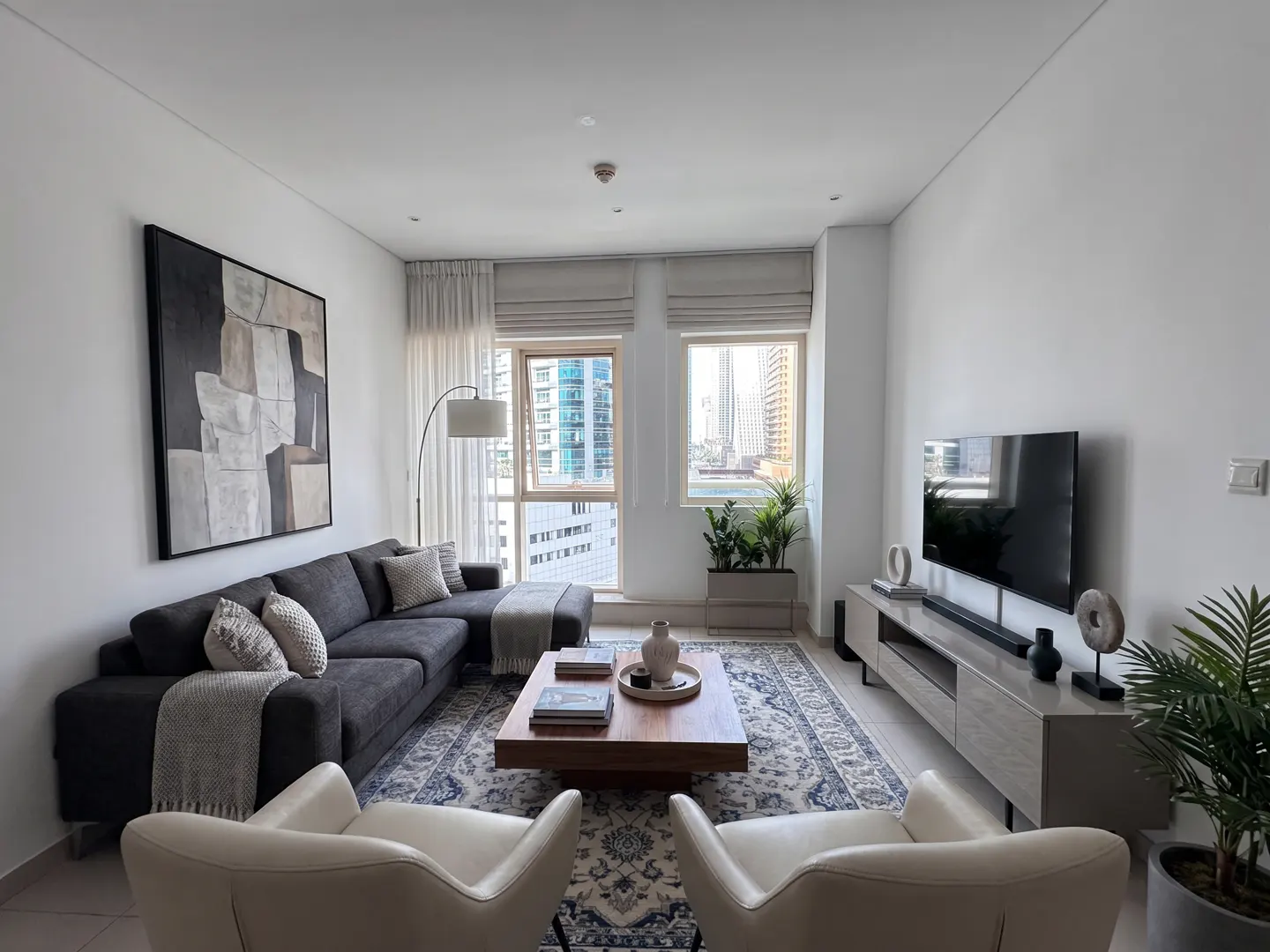 Living room with gray sectional sofa, two white chairs, wood coffee table, and a large abstract painting on the wall. Windows overlook a city skyline.