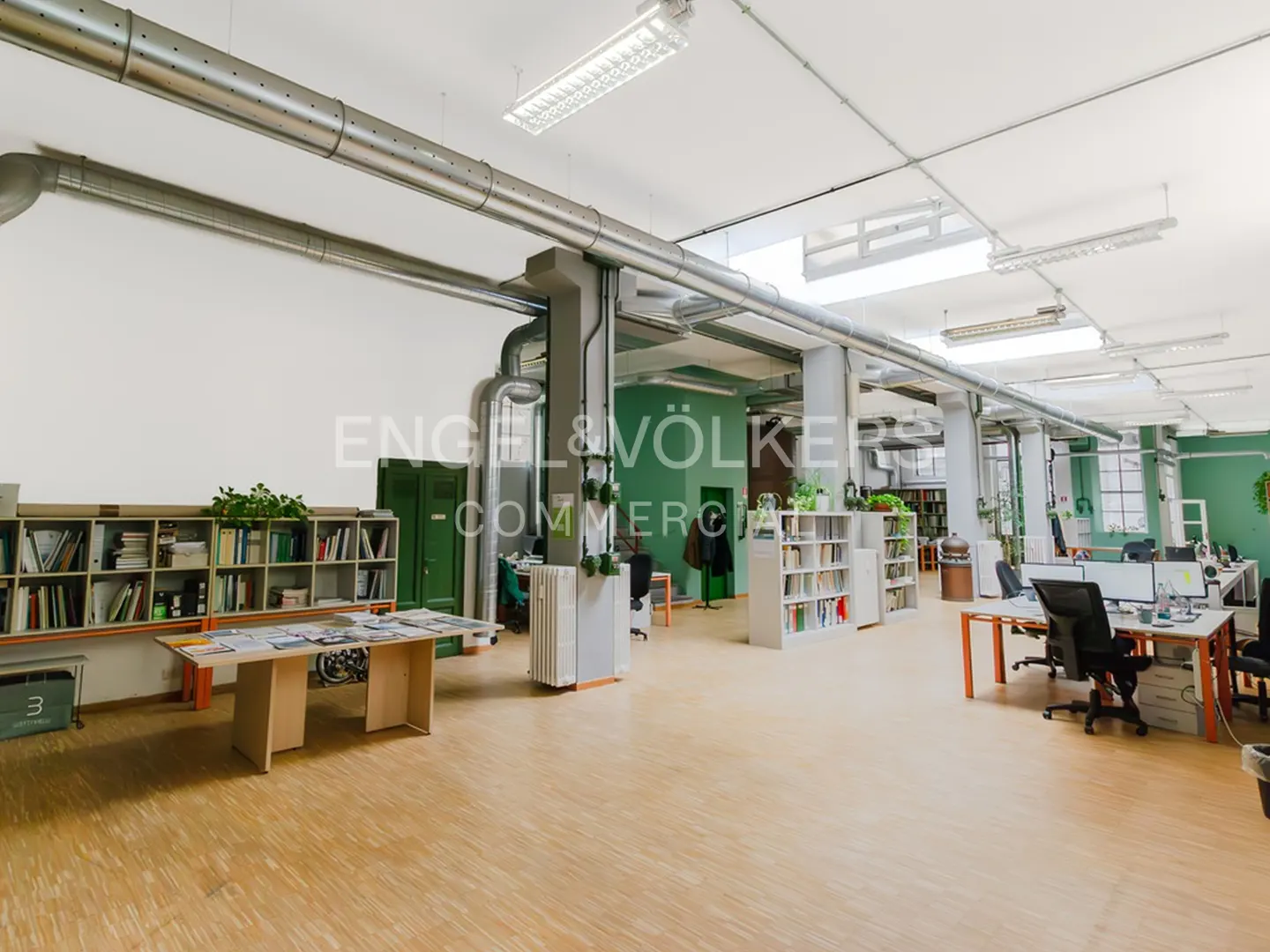 Bright, open office space with desks, chairs, bookshelves, and exposed ductwork. The walls are white and green. The floor is light wood.