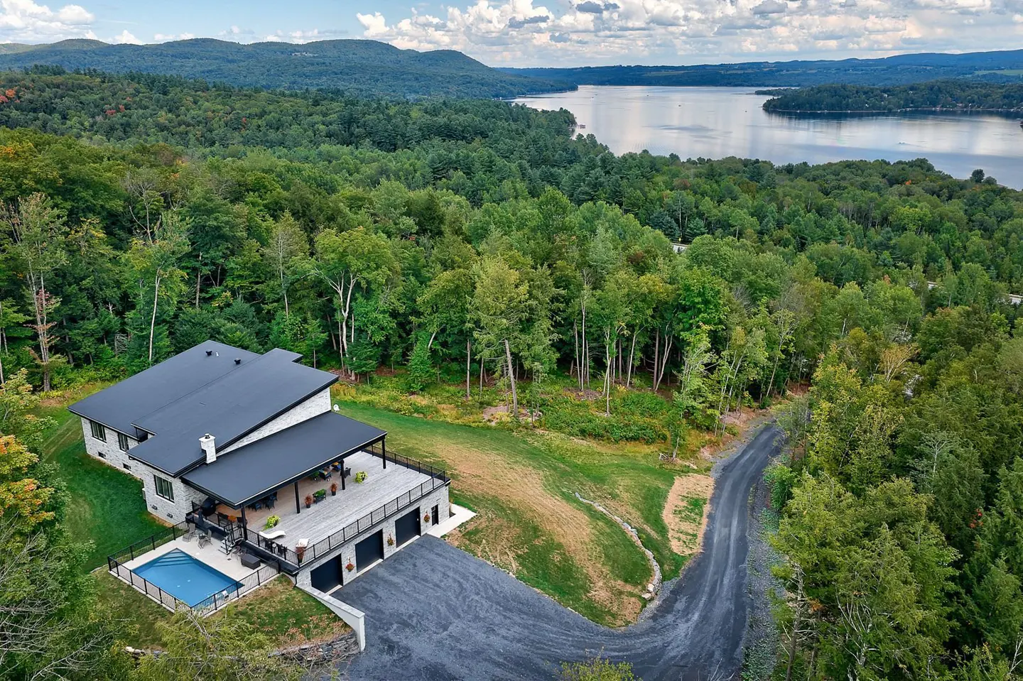 Aerial view of a modern stone house with a black roof, pool, and a lake view surrounded by lush green trees.