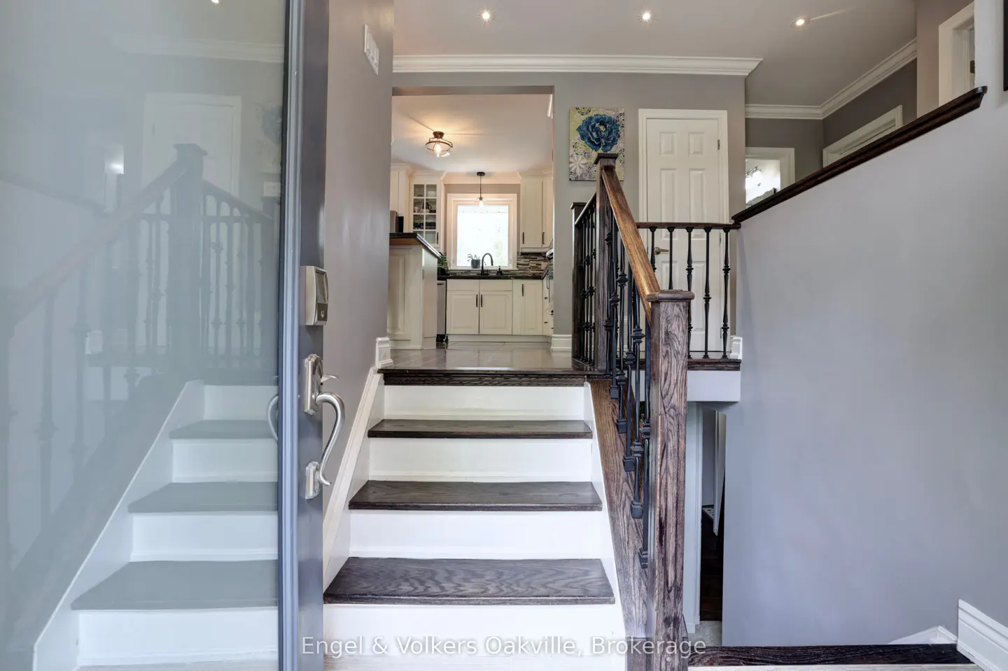 Open front door view of a home's foyer with stairs. White and dark wood stairs lead to the upper level. Kitchen visible in the background.