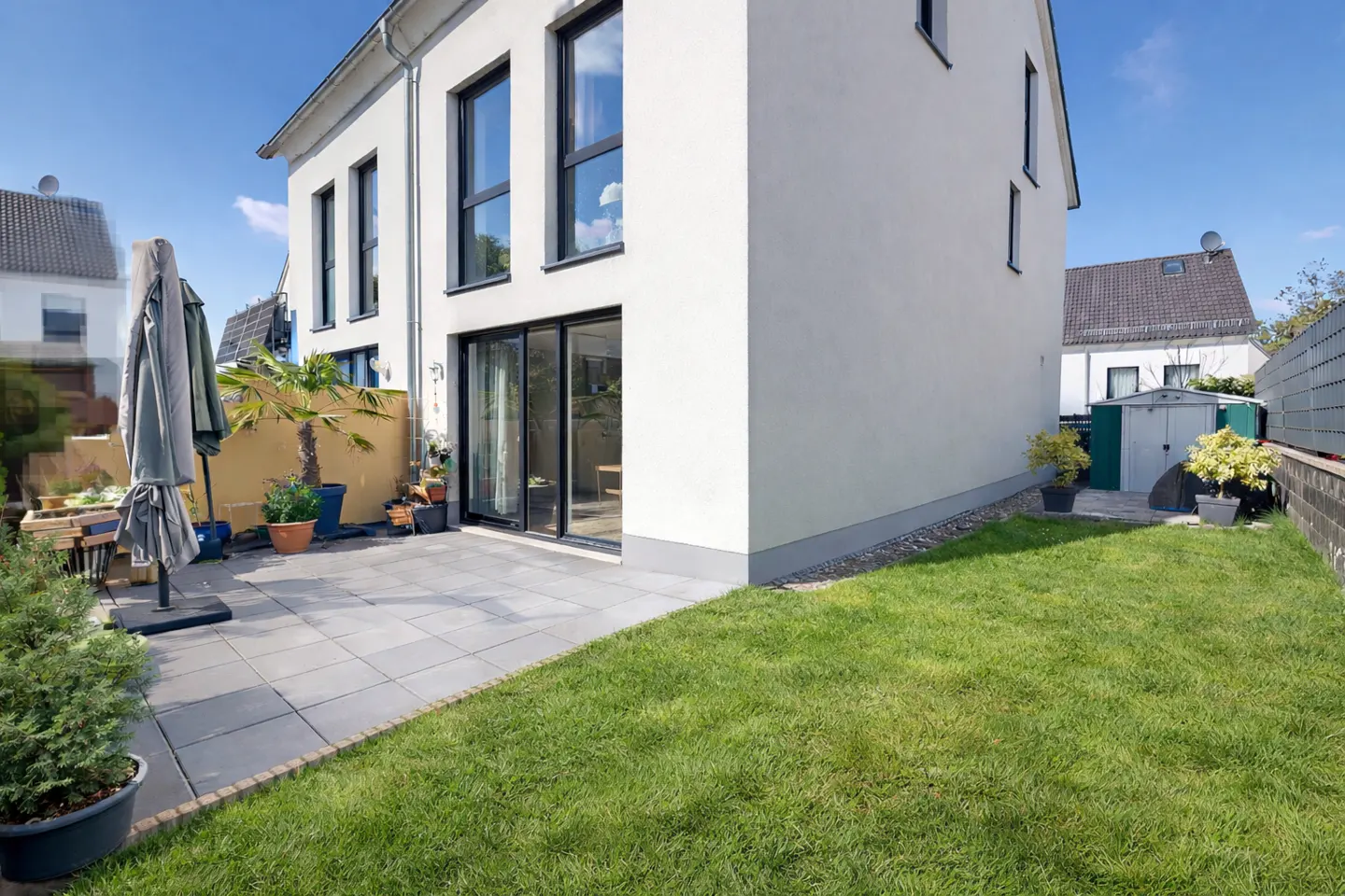 Backyard of a modern white house with a gray tiled patio, green lawn, and black framed windows under a blue sky.