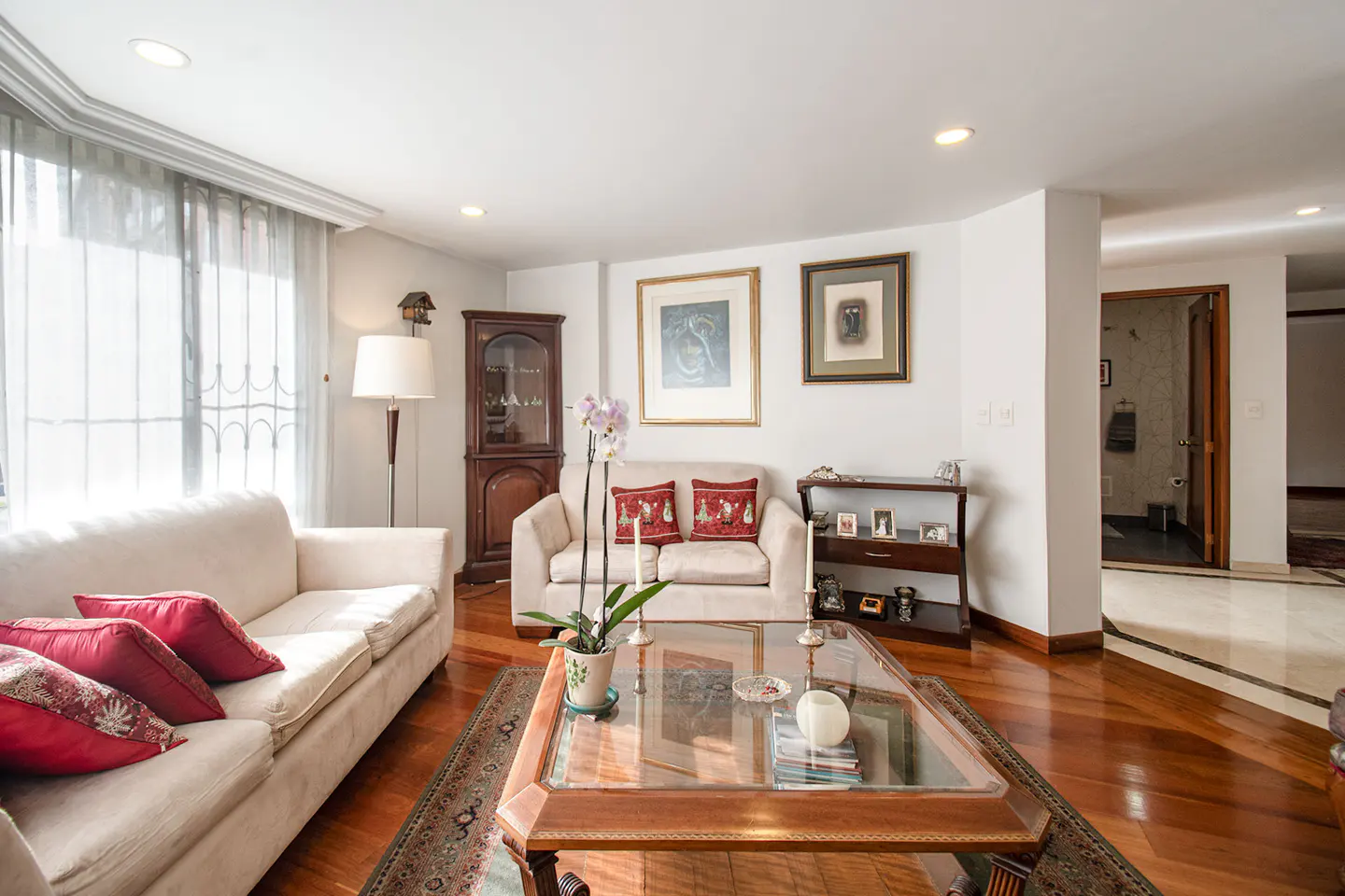Bright living room with beige sofas, red accent pillows, a glass coffee table, and hardwood floors.