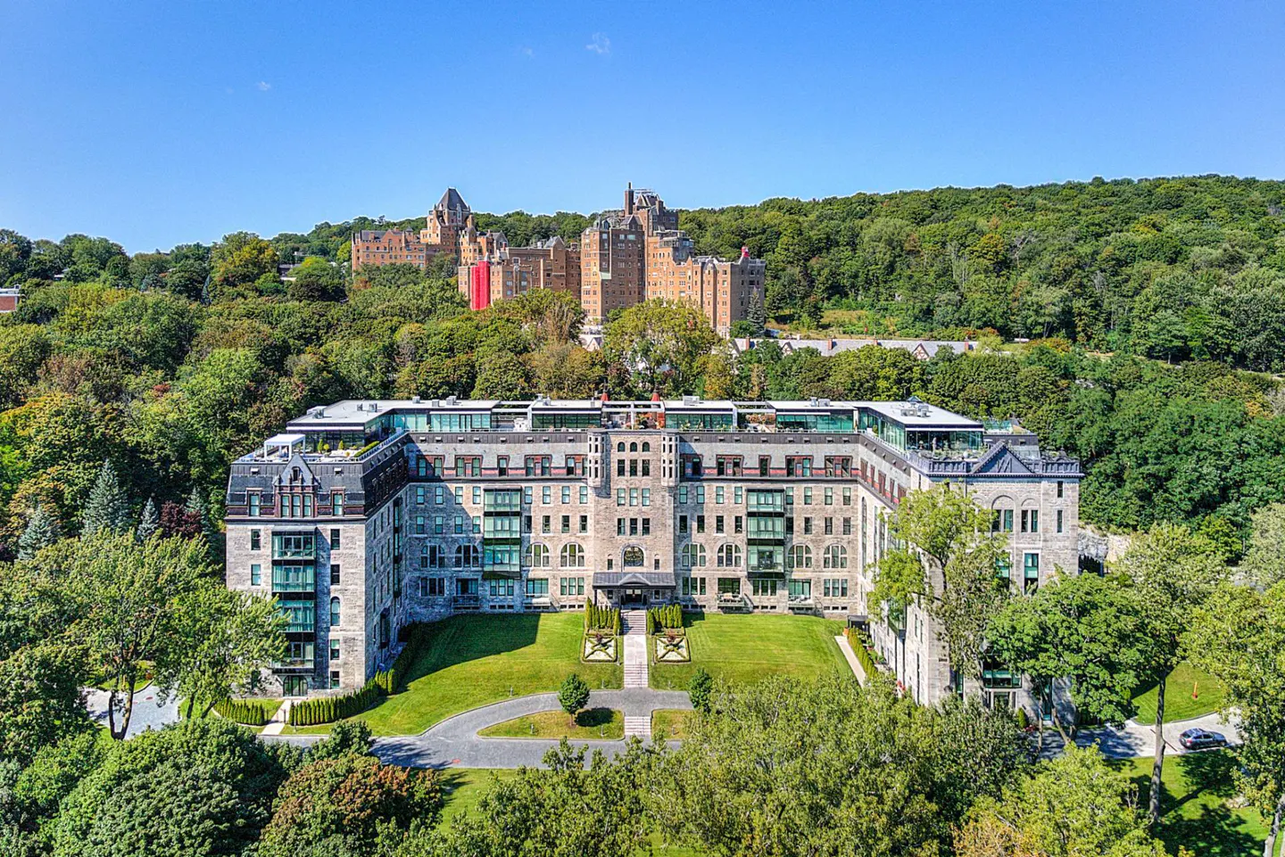 Aerial view of a large stone apartment building with green lawns, trees, and a castle in the background on a sunny day.