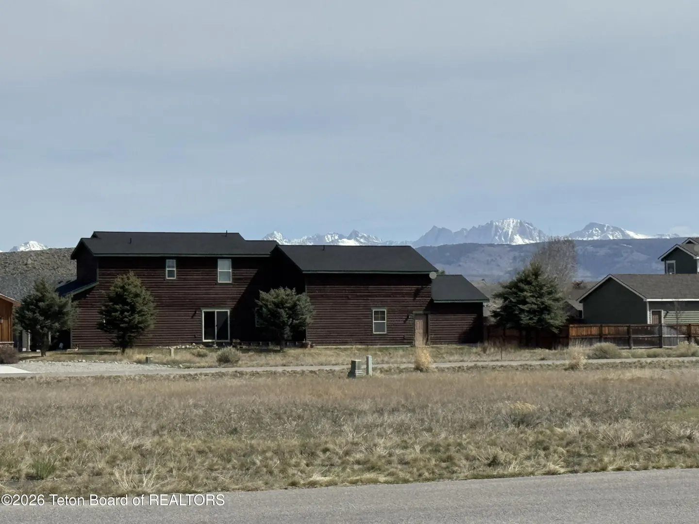 A two-story brown house with a black roof sits in front of snow-capped mountains under a blue sky.
