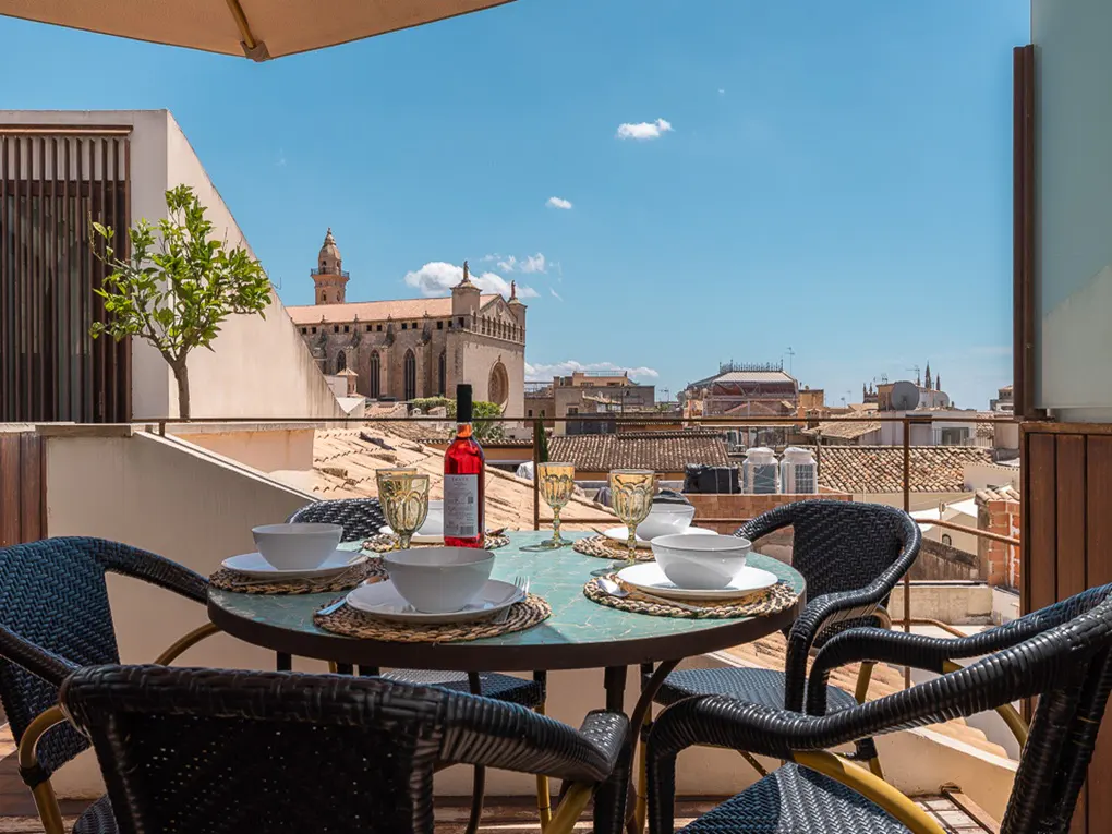 Outdoor dining area with a round table set for six, featuring a bottle of rosé wine, with a city view and blue sky in the background.
