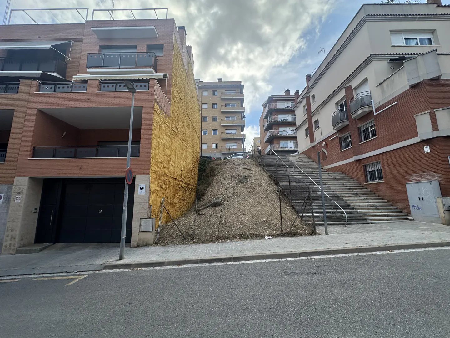 Street view of brick buildings with balconies, a dirt hill, and concrete stairs with metal railings.