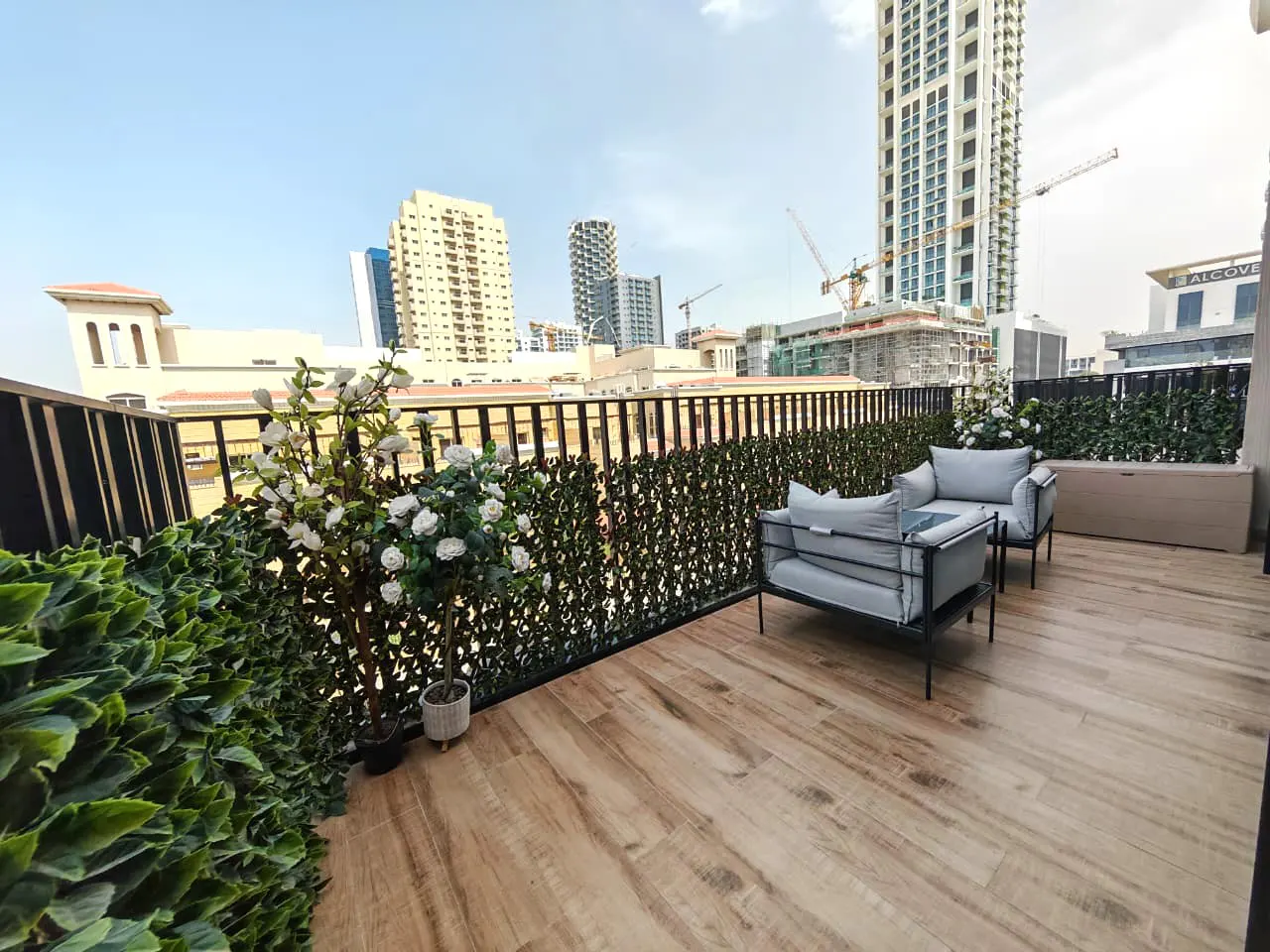 Balcony with wood-look tile, green privacy screen, and gray outdoor seating. City buildings are visible in the background.