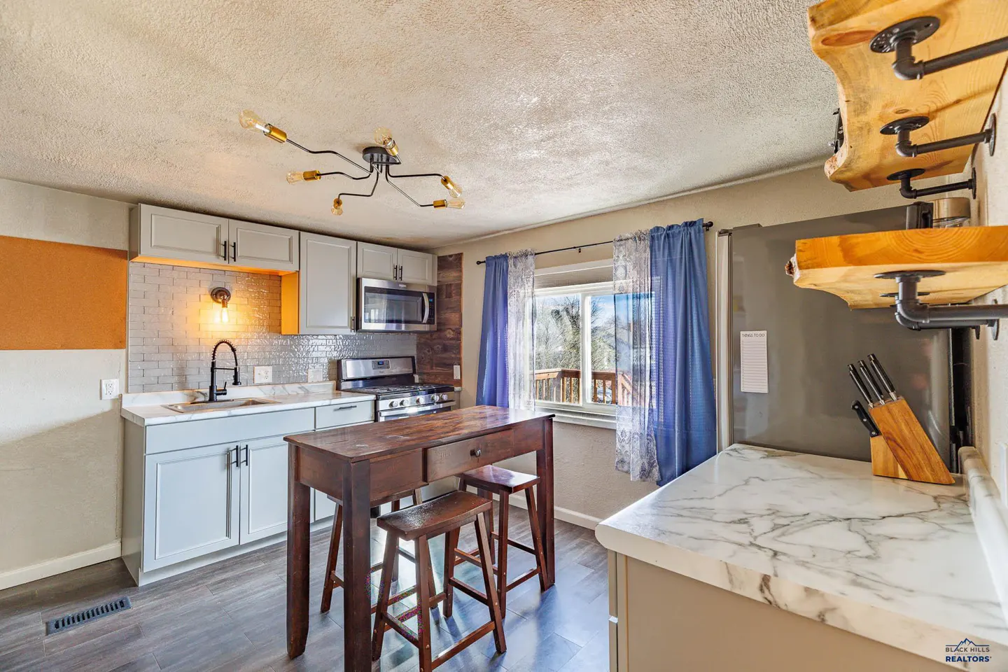 A kitchen with gray cabinets, marble countertops, and a dark wood island with stools. Blue curtains hang by a window.