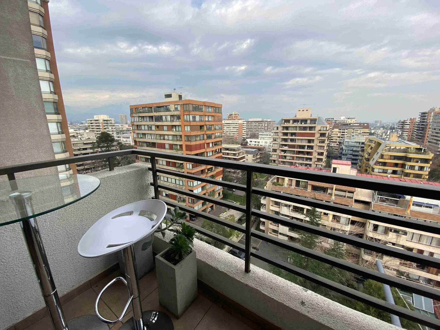 Balcony view of a city skyline with tall buildings under a cloudy sky. A glass table, white chair, and potted plant are on the balcony.
