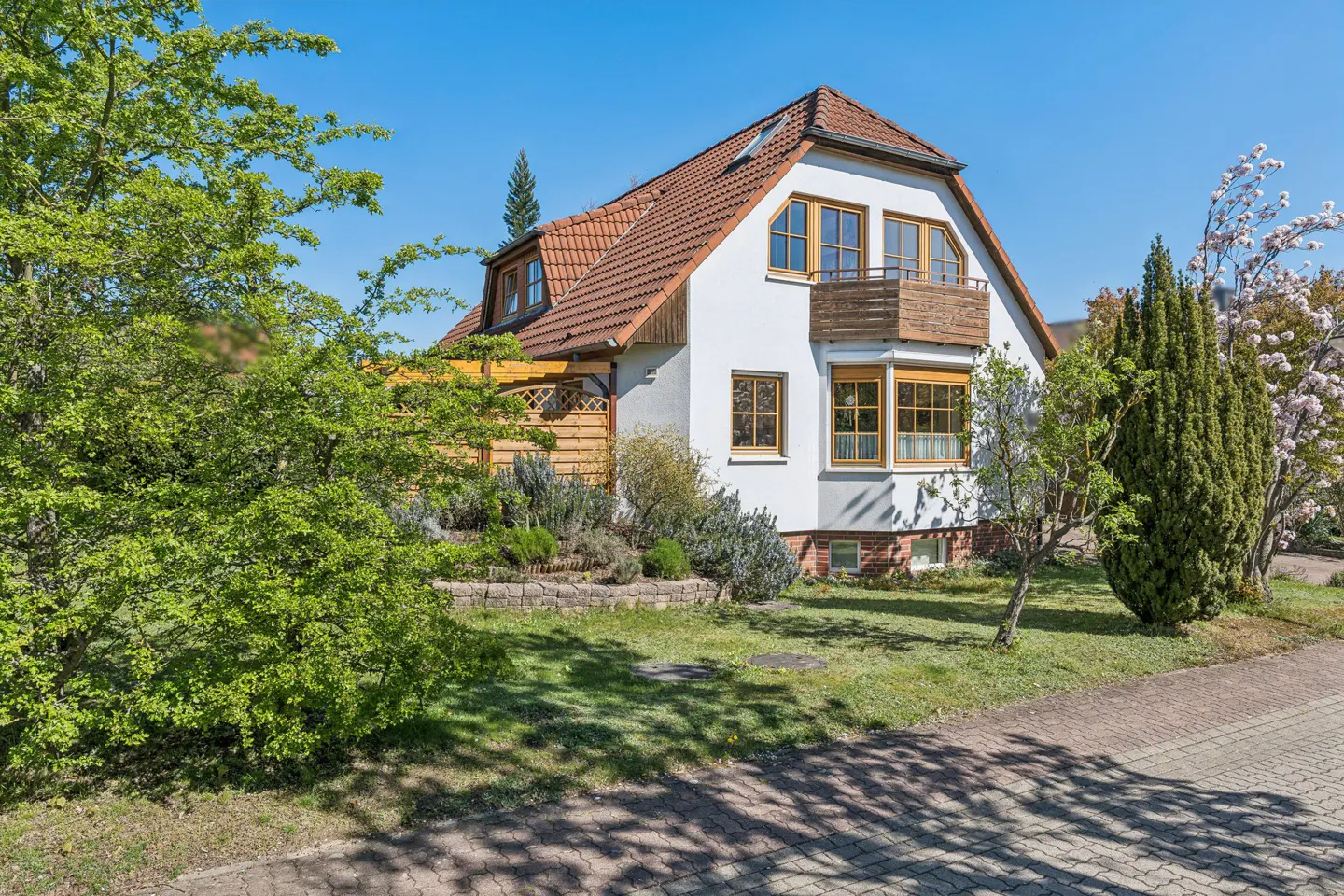 Two-story white house with a brown roof, wooden balcony, and green lawn on a sunny day.