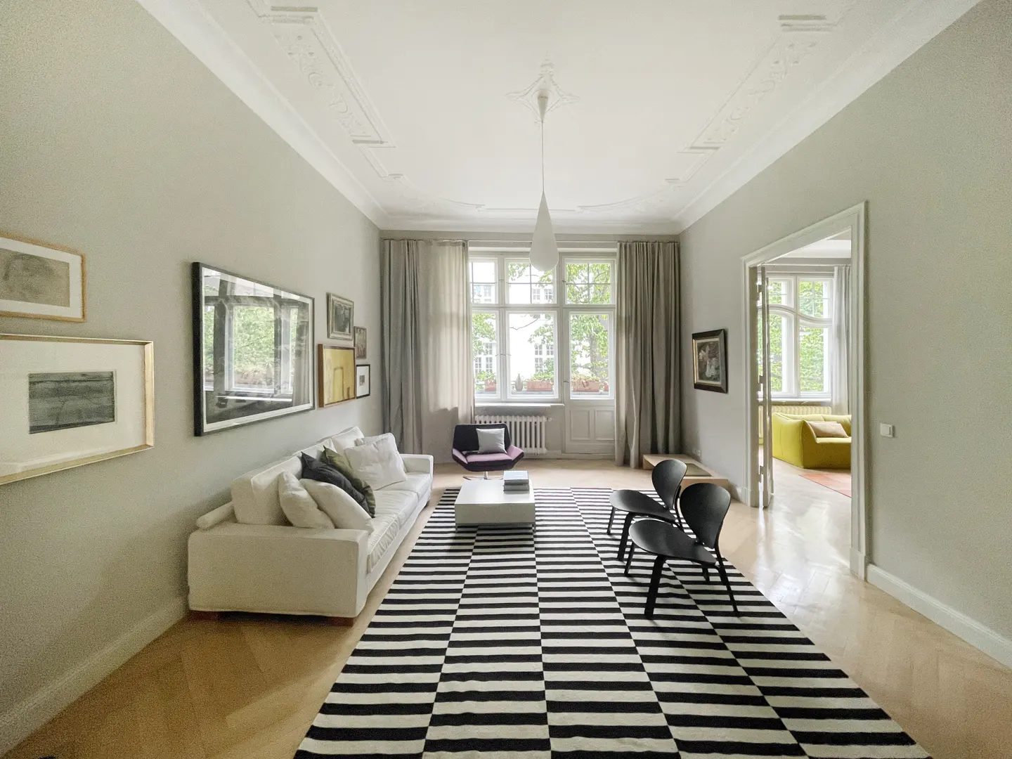 Bright living room with white sofa, black and white striped rug, two black chairs, and a view into a yellow-toned room.