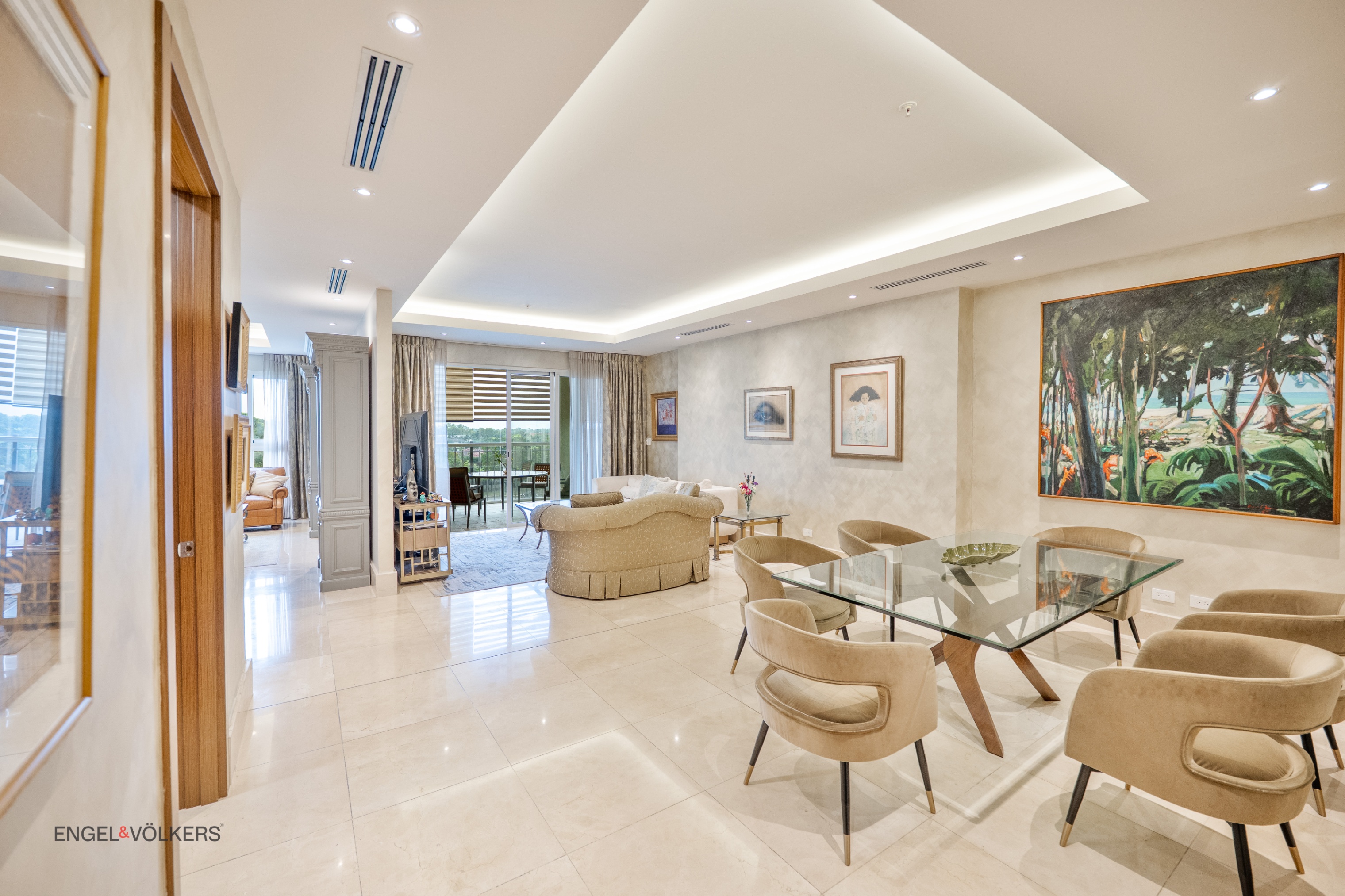 A bright, modern living and dining area with beige walls, marble floors, and a glass dining table with tan chairs. A large painting hangs on the wall.