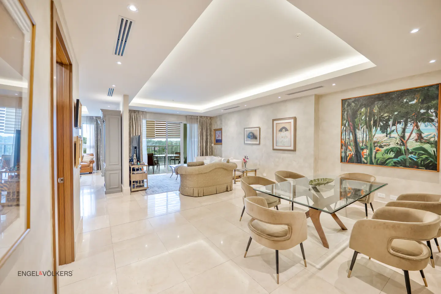 A bright, modern living and dining area with beige walls, marble floors, and a glass dining table with tan chairs. A large painting hangs on the wall.