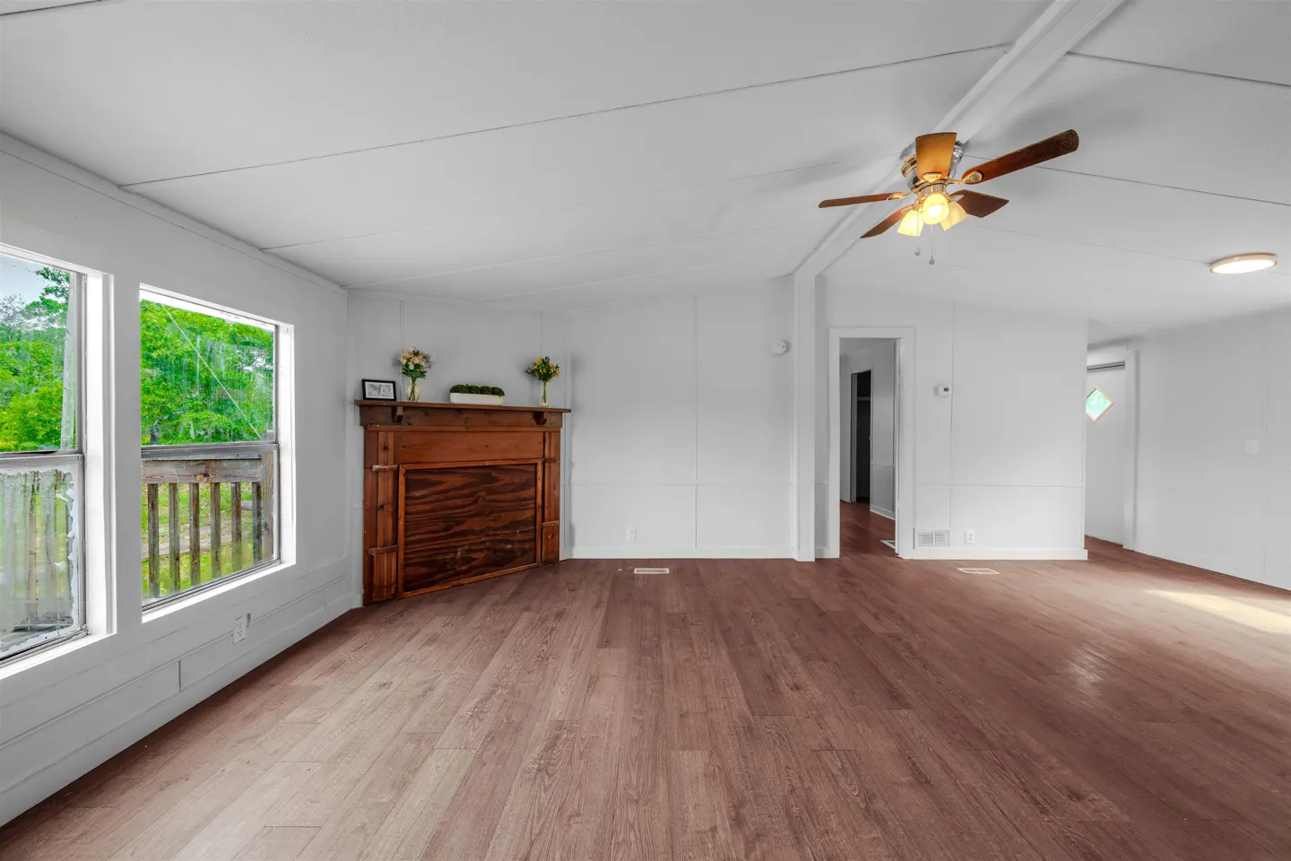 Bright living room with wood floors, white walls, and a brown fireplace. Windows show green trees outside. A ceiling fan hangs above.