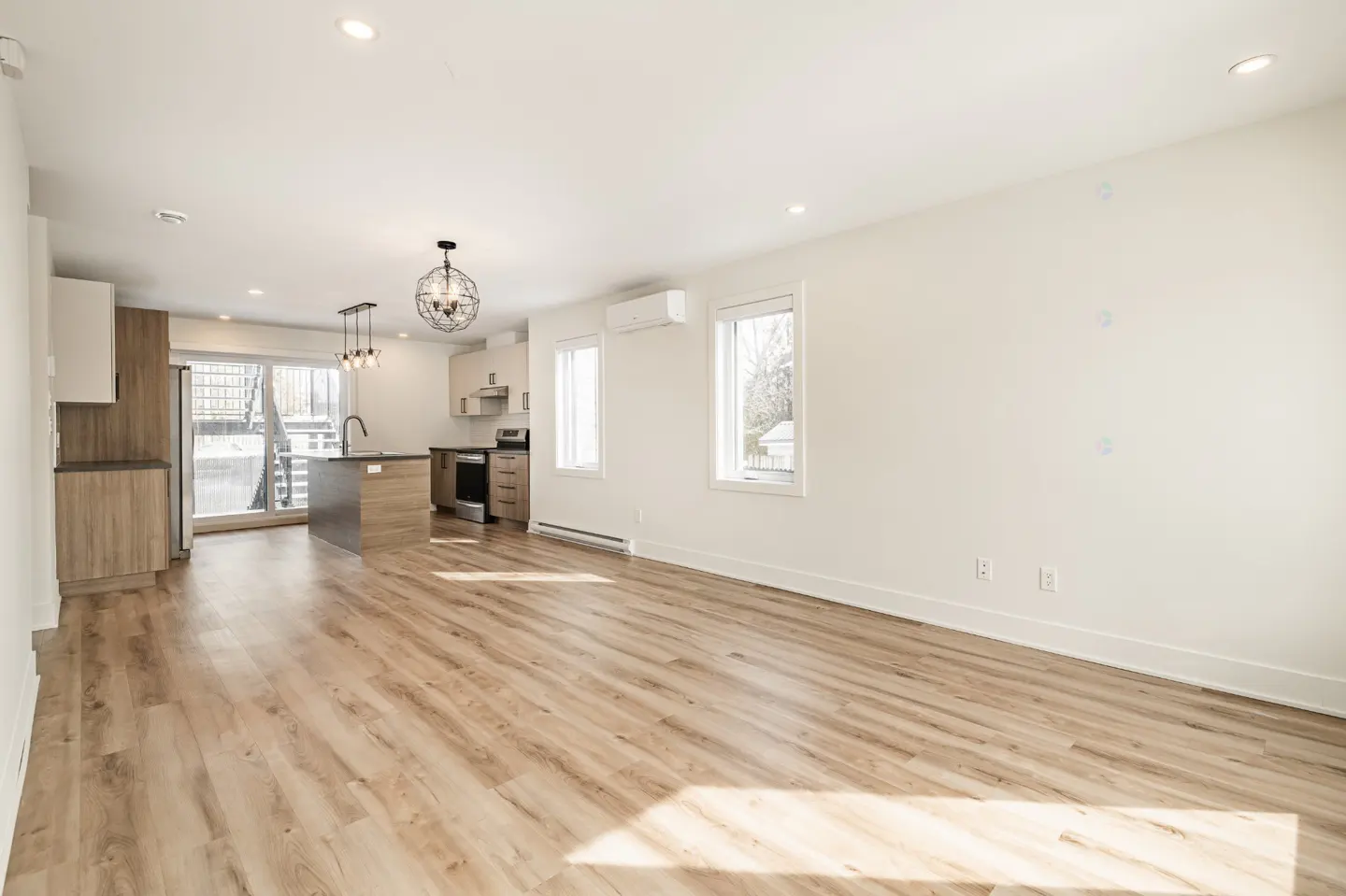 Bright, open-concept apartment with wood floors, white walls, and a modern kitchen with stainless steel appliances. Windows let in natural light.