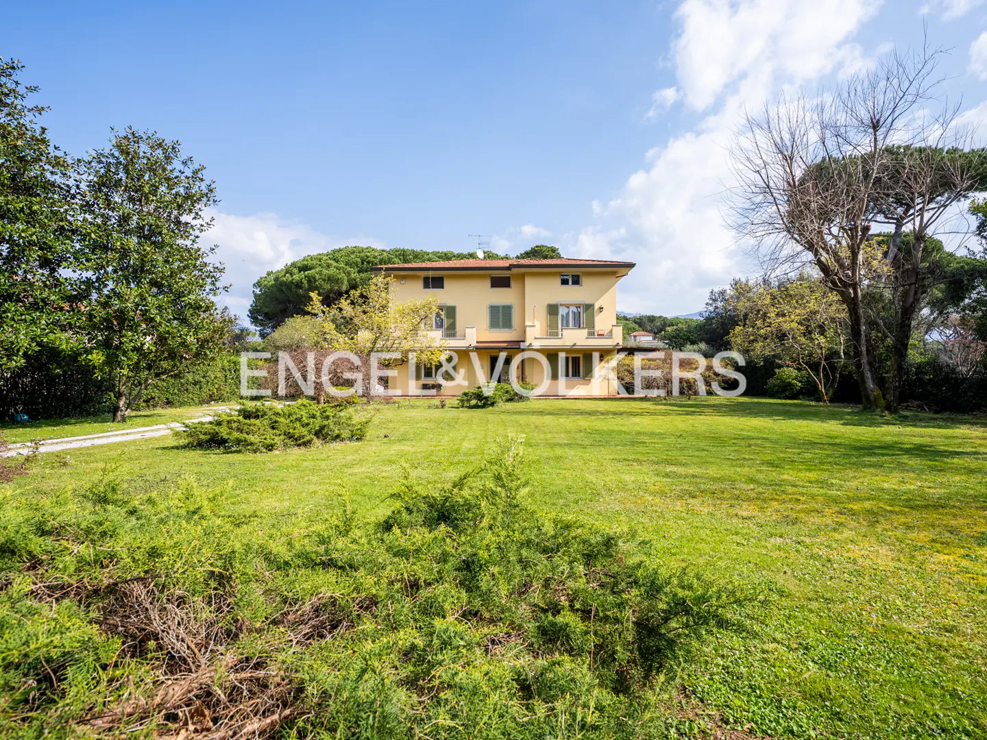 Two-story yellow house with green shutters, surrounded by a large green lawn and trees under a blue sky.
