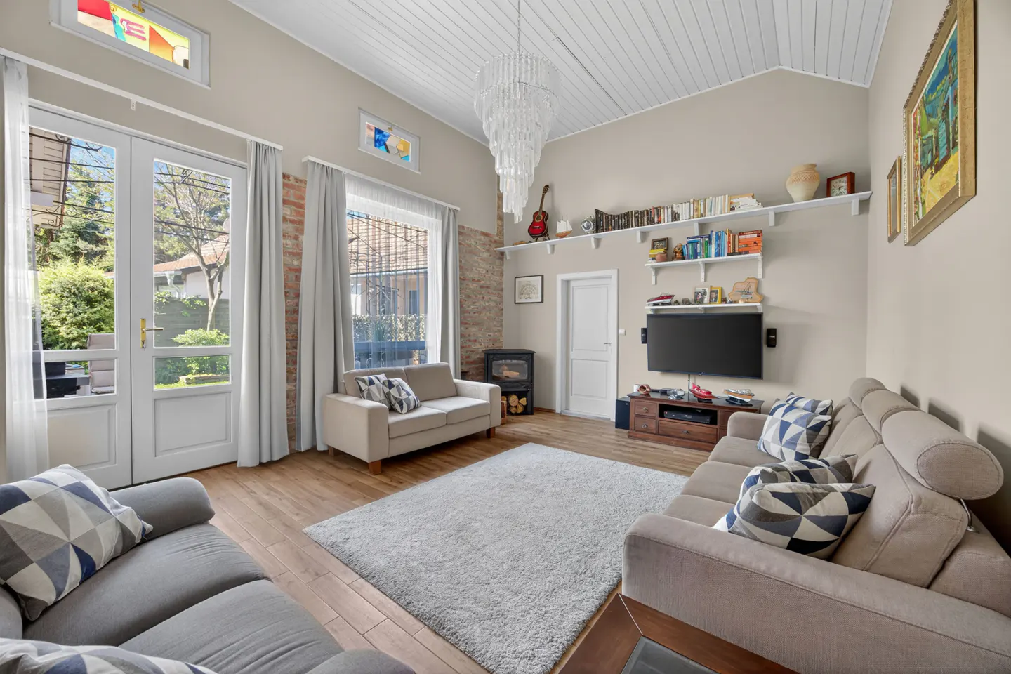 Living room with gray sofas, wood floors, and a white chandelier. Shelves with books and a TV are on the wall. French doors lead to a garden.