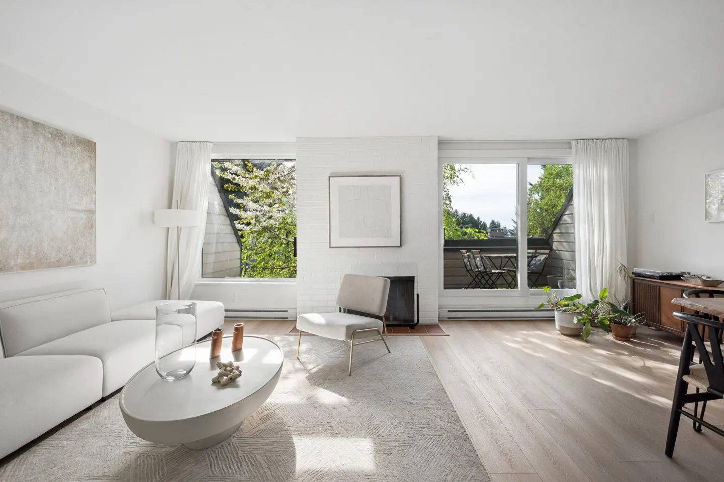 Bright living room with white sofa, oval coffee table, and chair. Large windows offer views of greenery and a balcony.