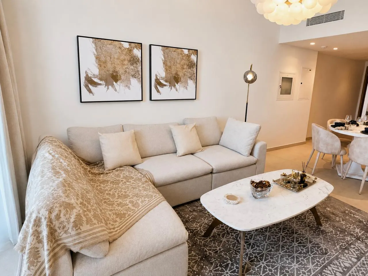 Living room with a beige sectional sofa, marble coffee table, and abstract art. Dining area visible in the background.