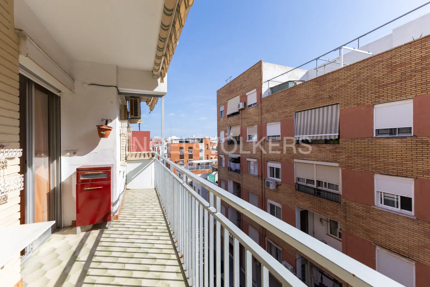 View from a sunny apartment balcony overlooking a neighboring brick residential building under a clear blue sky.