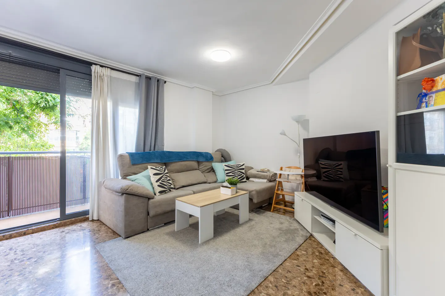Bright living room with a gray sofa, a light wood coffee table, and a large TV. Balcony doors let in natural light.
