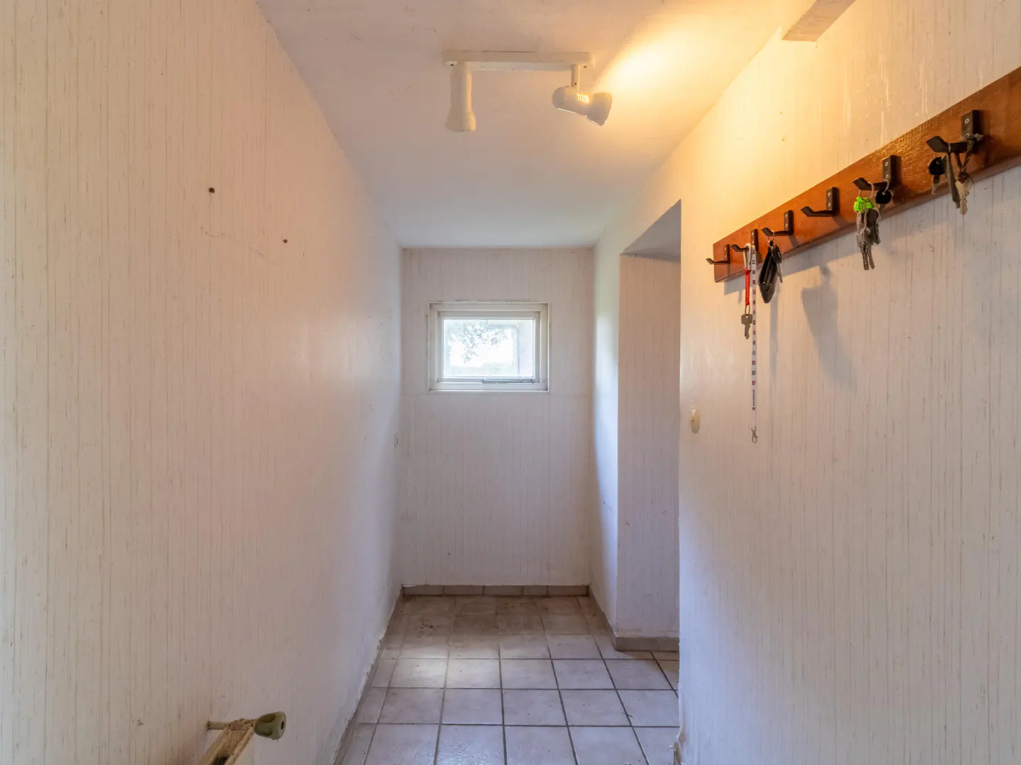 A narrow hallway with white walls, a tiled floor, and a window at the end. A coat rack with keys hangs on the right wall.