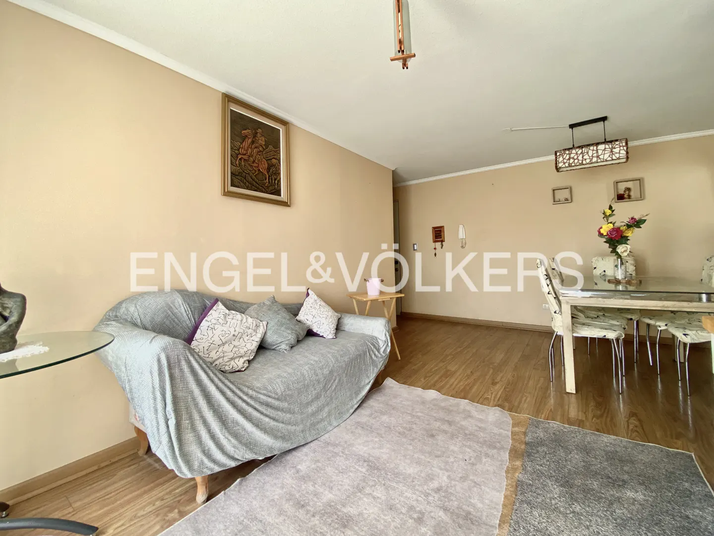 Living room with beige walls, wood floors, and a gray sofa with pillows. A dining table with chairs is in the background. Artwork hangs on the walls.