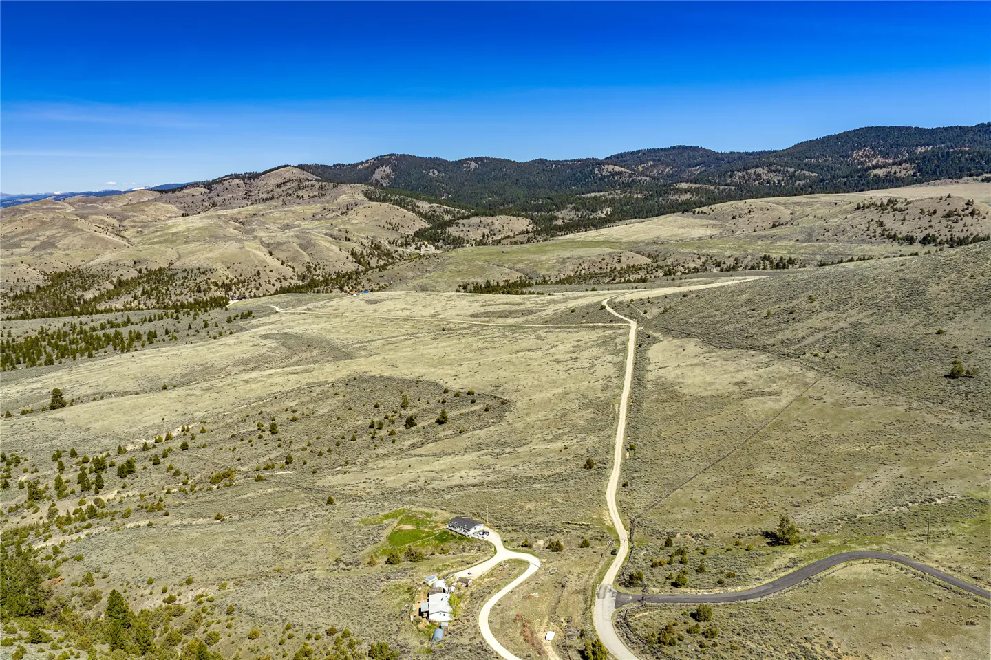 Aerial view of a rural property with a long driveway leading to a house, set against a backdrop of rolling hills and a clear blue sky.