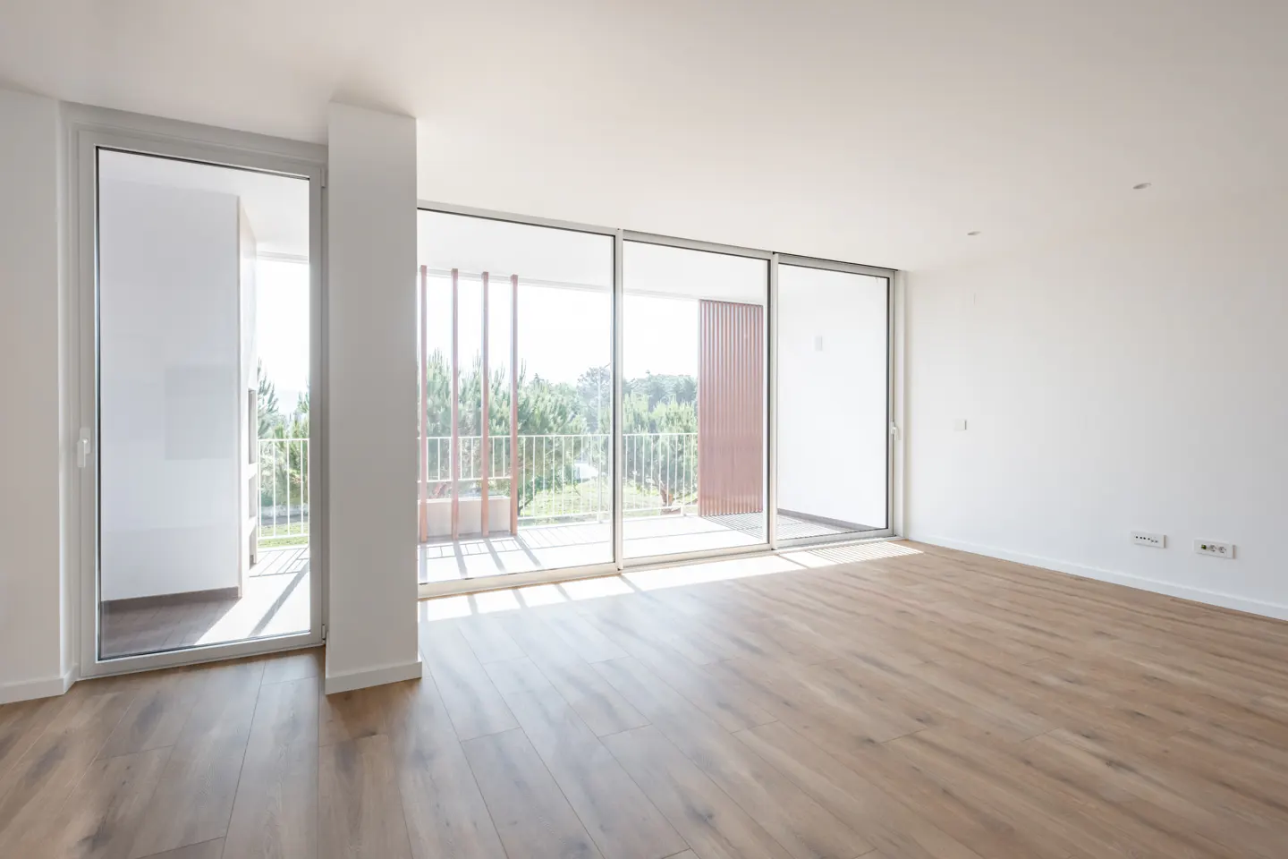 Bright, empty room with wood floors and white walls. Large glass doors lead to a balcony with a view of trees.