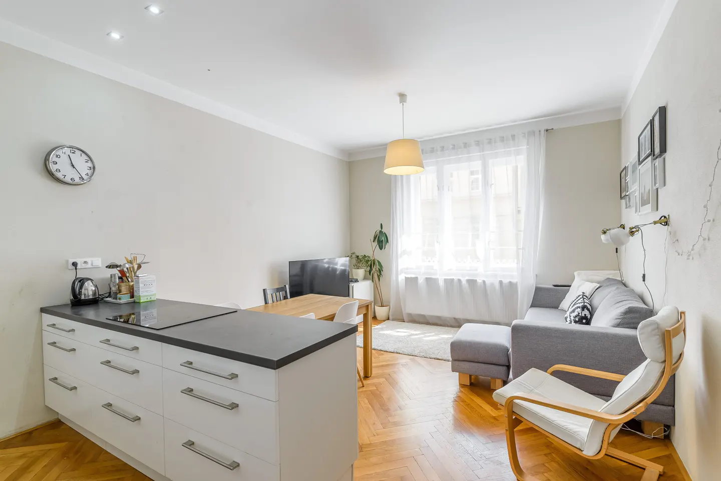 Bright apartment interior with a gray sofa, white chair, and kitchen island with a black countertop. Hardwood floors and a window with sheer curtains.