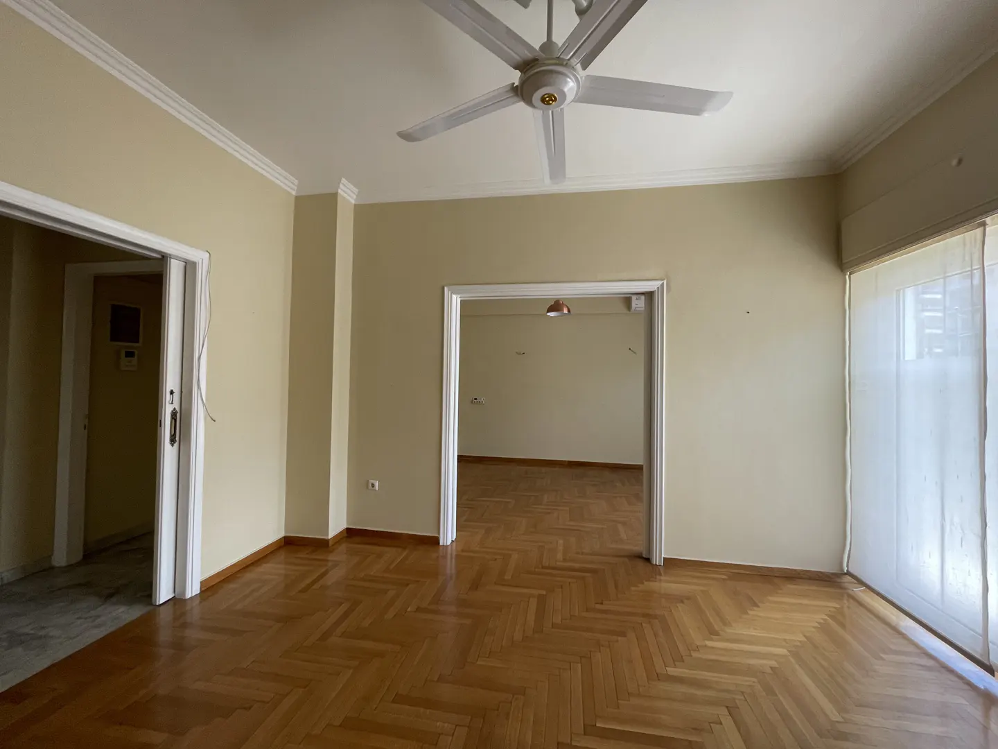 Empty room with herringbone wood floors, beige walls, and white trim. A ceiling fan hangs above, and doorways lead to other rooms. Natural light filters through window blinds.