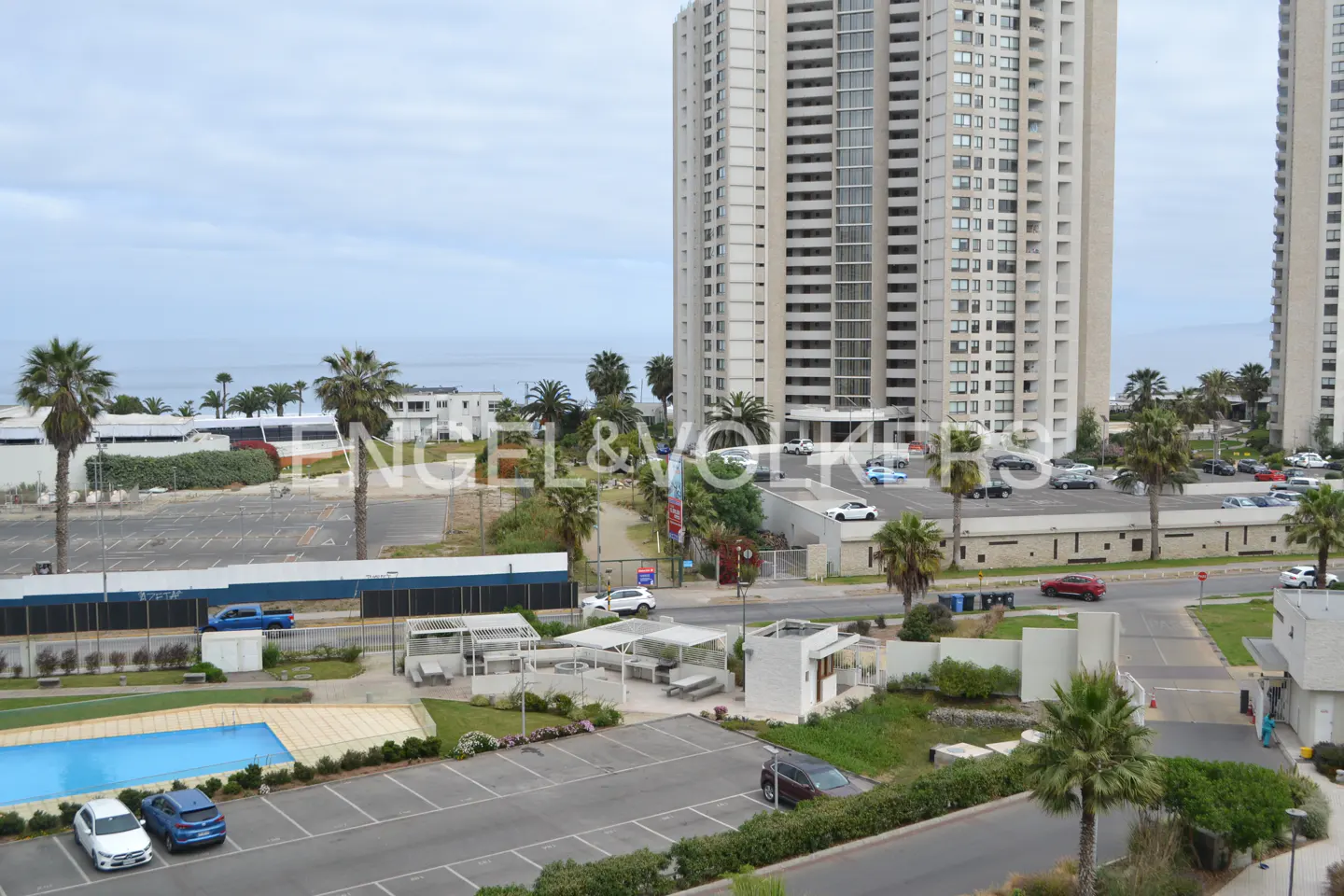 View of a tall apartment building near the ocean, with a pool, parking lot, and palm trees.