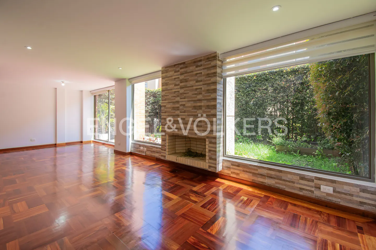 Bright living room with parquet floors, a stone fireplace, and a large window overlooking a green garden.