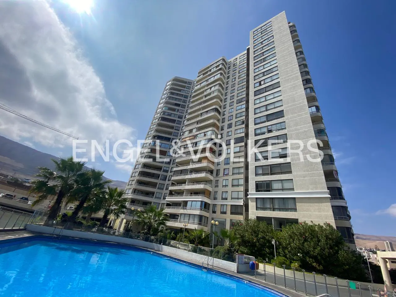 Exterior shot of a tall, modern apartment building with balconies, a blue swimming pool, palm trees, and a clear blue sky.