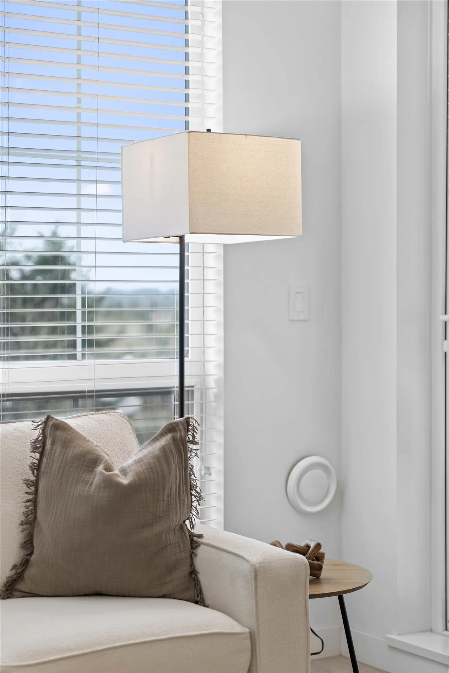 A beige sofa with a brown pillow sits next to a modern floor lamp and a small wooden side table by a window with blinds.