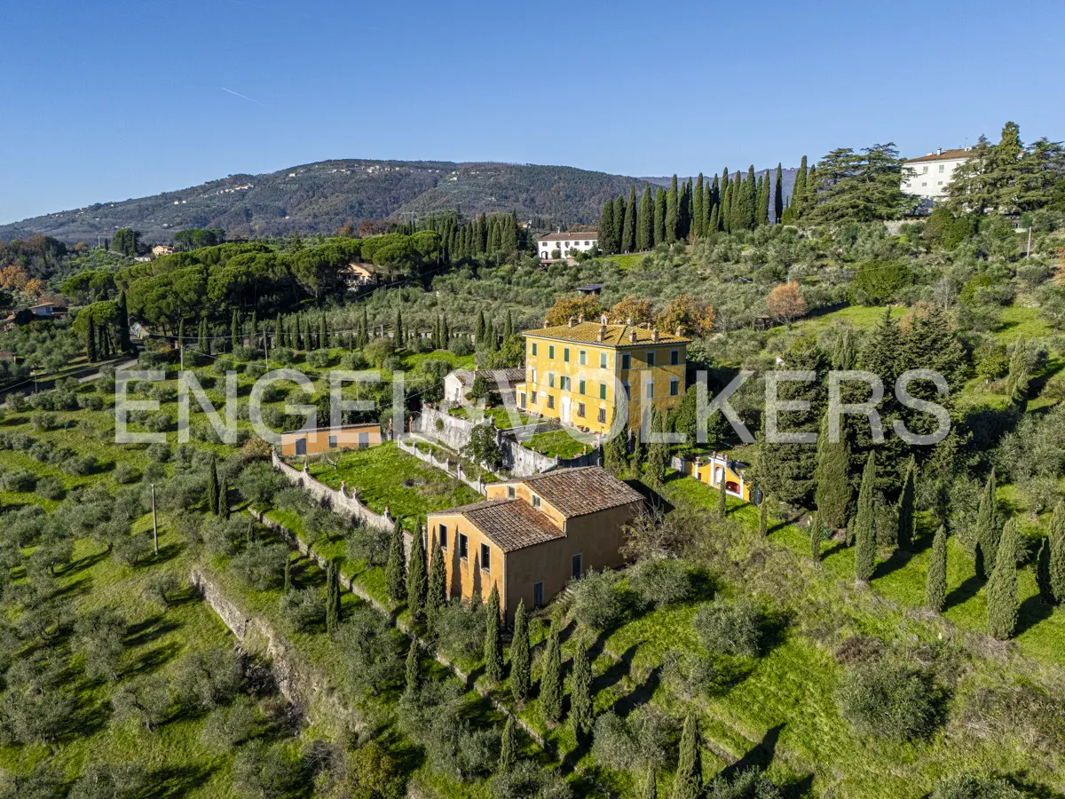 Aerial view of a yellow villa with a red tile roof, surrounded by green trees and rolling hills under a blue sky.
