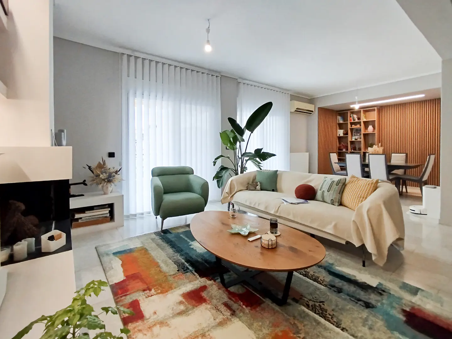 Bright living room with a green armchair, beige sofa, and wooden coffee table on a colorful rug. A fireplace and dining area are visible.
