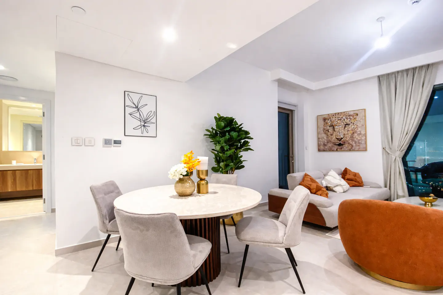 Bright living room with a round dining table, grey chairs, and a sofa with orange pillows. A large window shows a city view.