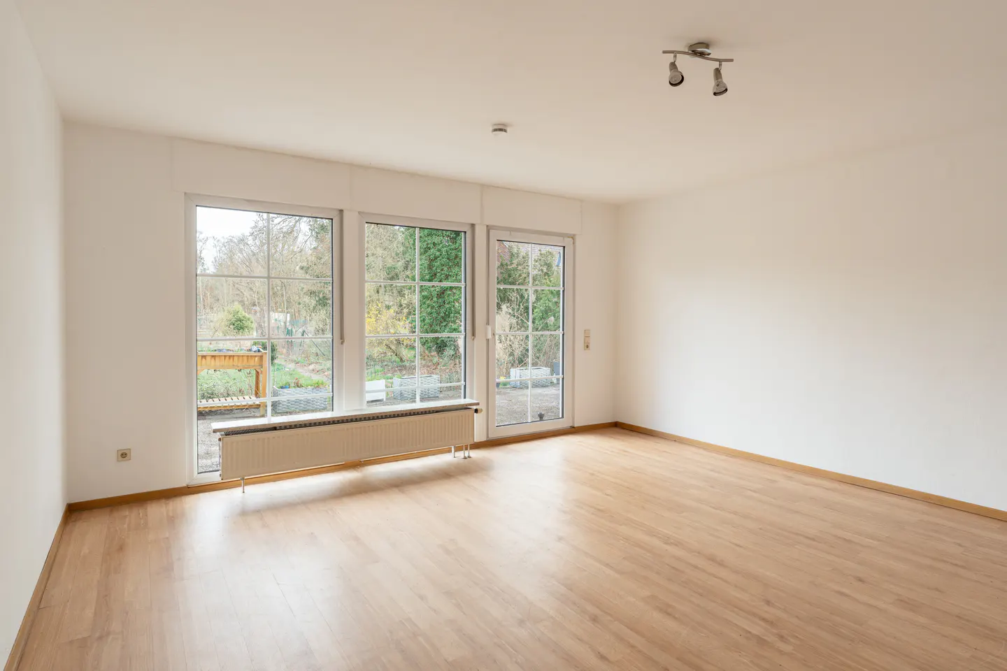 Bright, empty room with wood floors, white walls, and three large windows overlooking a garden. A radiator sits beneath the windows.