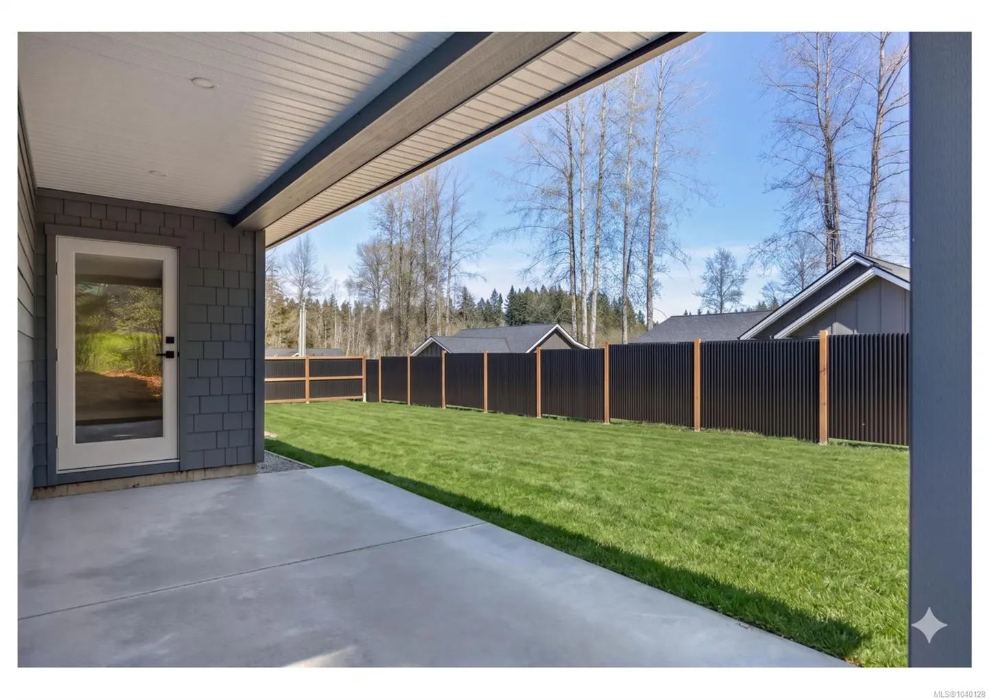 A covered concrete patio overlooks a green backyard enclosed by a modern black corrugated metal fence under a blue sky.