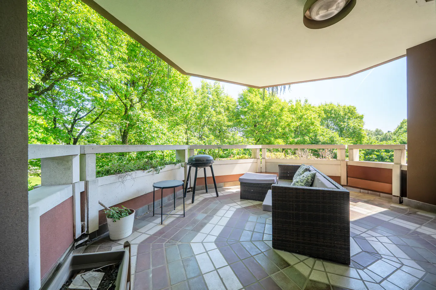 Covered balcony with outdoor furniture, grill, and tiled floor. Green trees visible beyond the railing.