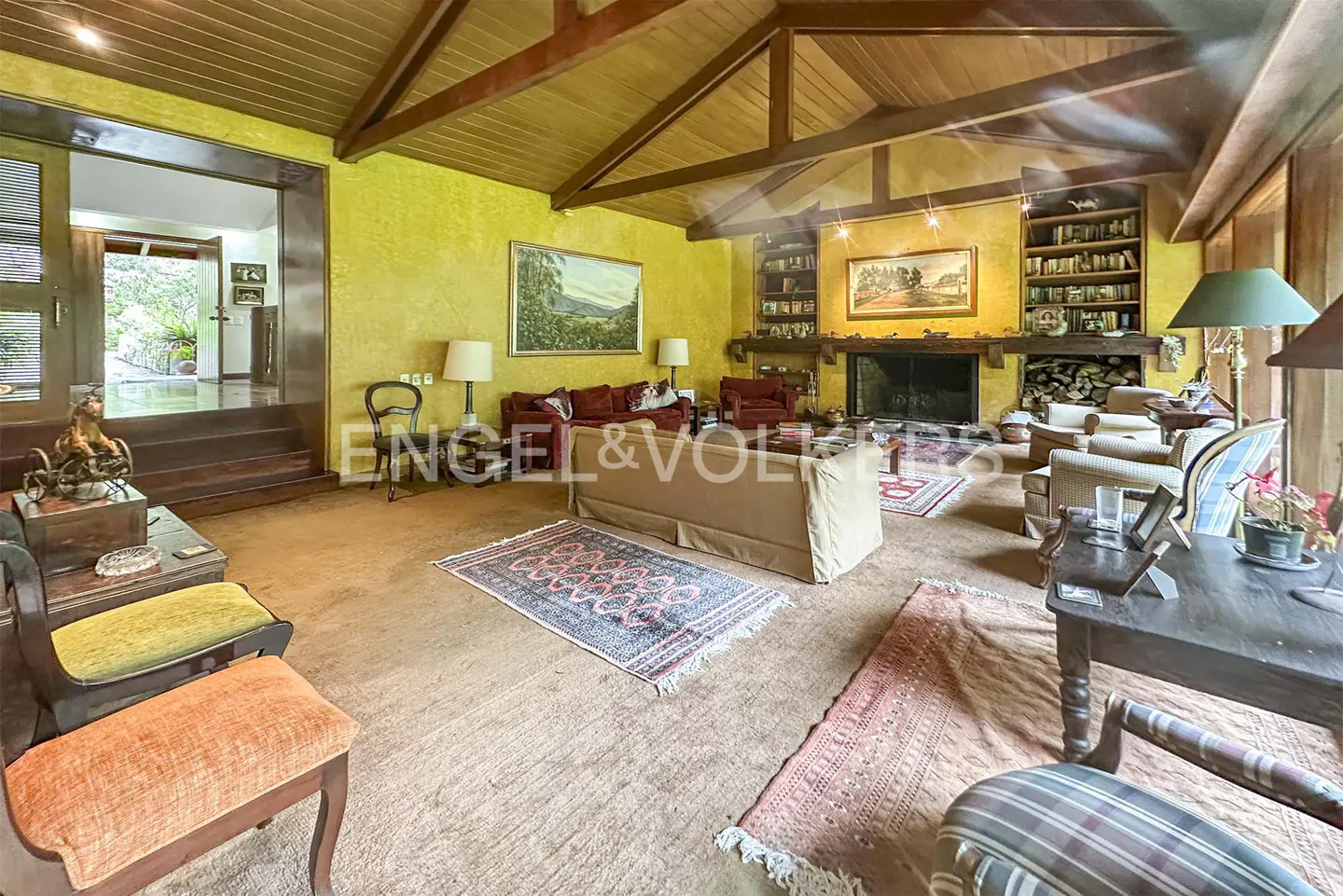 A spacious living room with a wooden ceiling, yellow walls, and a fireplace. There are sofas, chairs, rugs, and bookshelves.