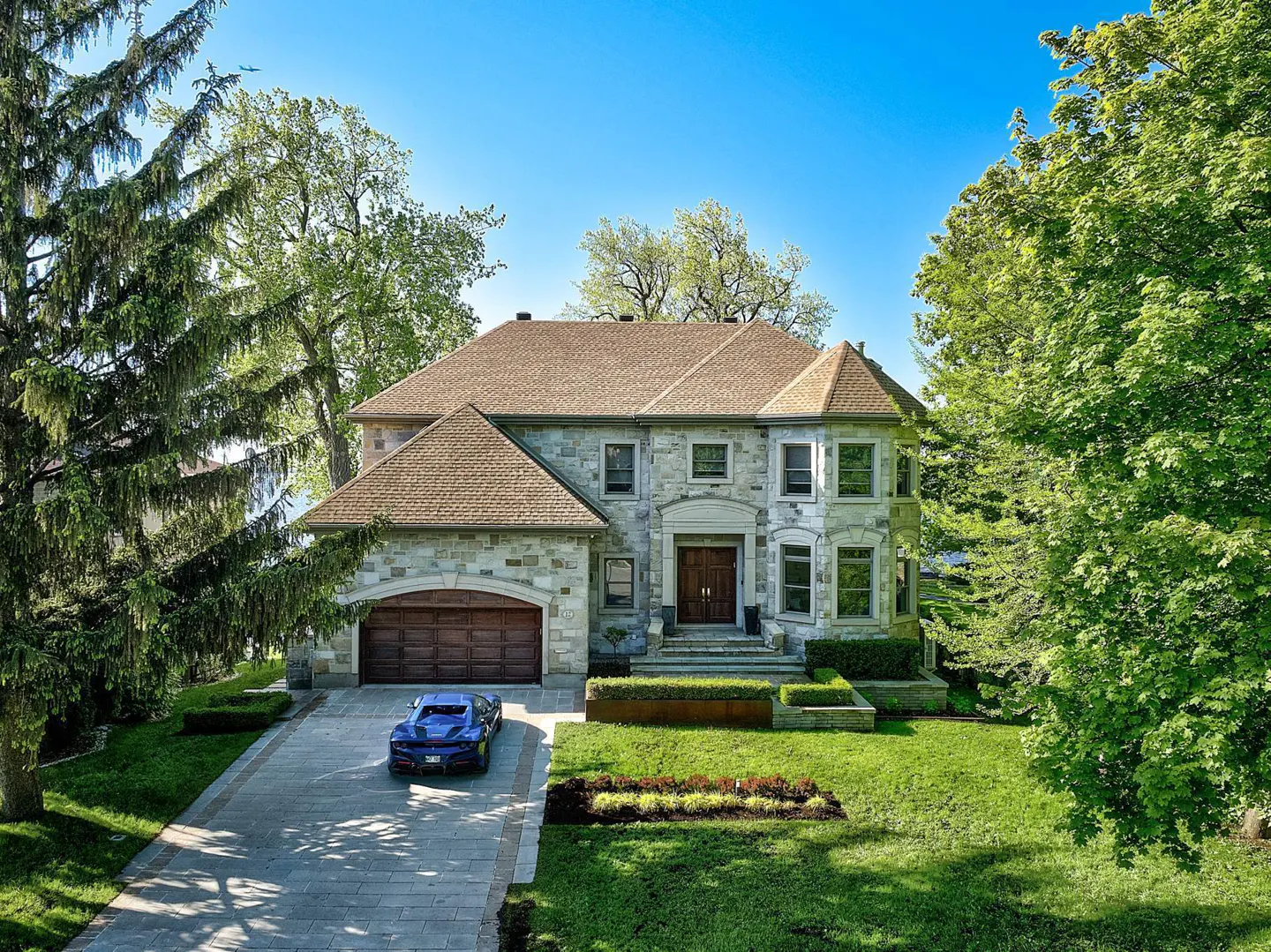 Two-story stone house with a brown roof, green lawn, and a blue sports car in the driveway.